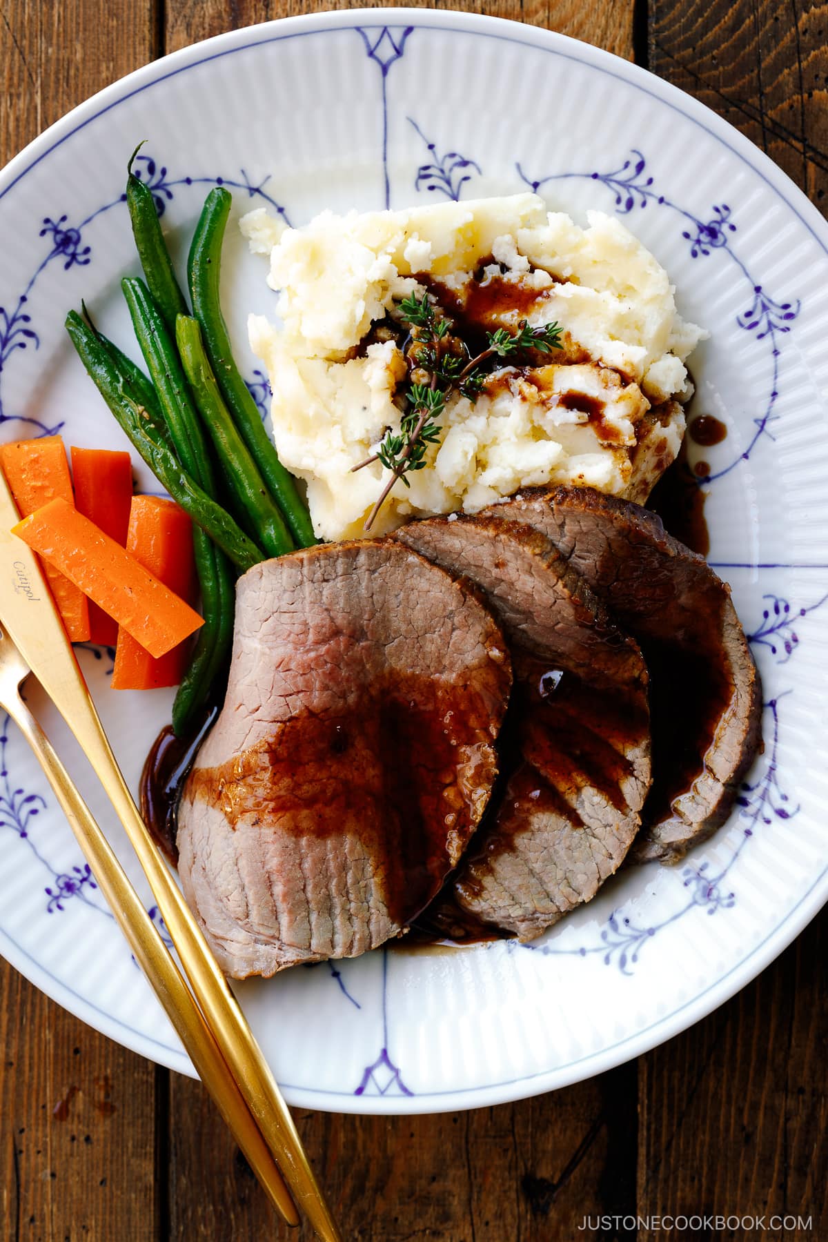 A white and blue plate containing roast beef with gravy, mashed potatoes, green beans, and carrot.