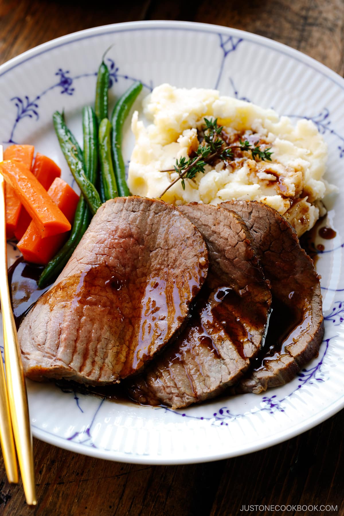 A white and blue plate containing roast beef with gravy, mashed potatoes, green beans, and carrot.