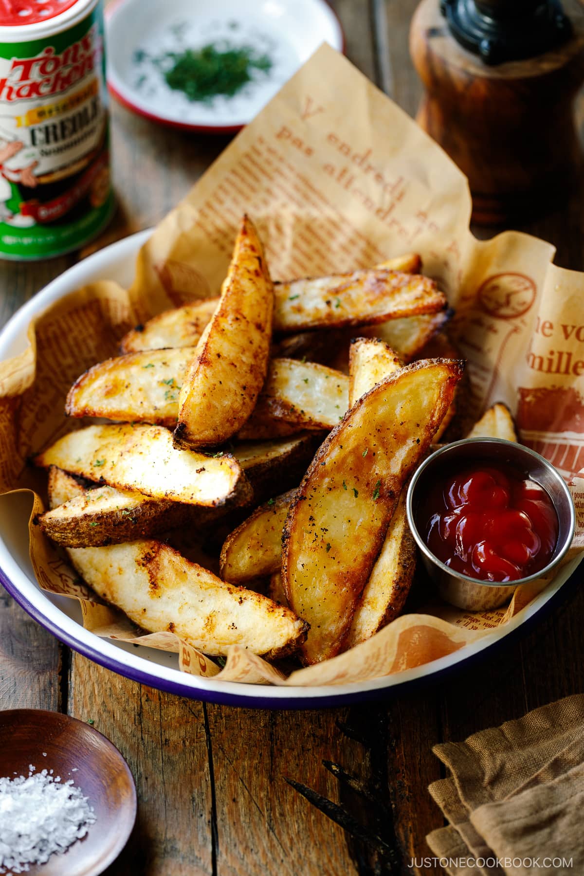 A white and blue enamel bowl containing Crispy Baked Potato Wedges with ketchu in a mini container.