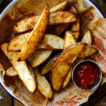 A white and blue enamel bowl containing Crispy Baked Potato Wedges with ketchu in a mini container.