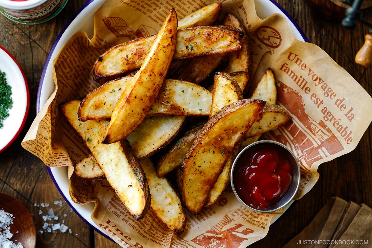 A white and blue enamel bowl containing Crispy Baked Potato Wedges with ketchu in a mini container.