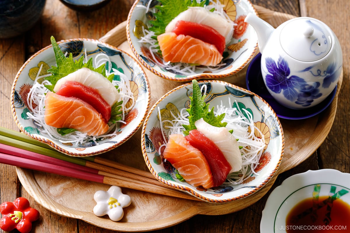 Multiple bowls containing sashimi grade fish, including salmon, tuna, and kanpachi, garnished with shredded daikon, shiso leaves, and wasabi.