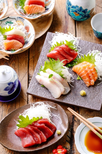 Multiple plates containing sashimi grade fish, including salmon, tuna, and kanpachi, garnished with shredded daikon, shiso leaves, and wasabi.