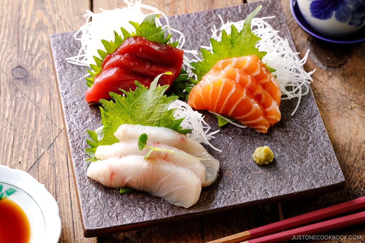 A plate containing sashimi grade salmon, kanpachi, and tuna, garnished with shredded daikon, shiso leaves, and wasabi.