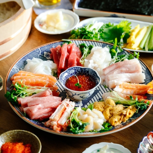 A temaki hand roll sushi spread on the dining table, consisting of plates and bowls containing sushi rice, sashimi, vegetable fillings, and nori seaweed.