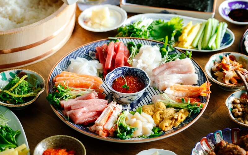 A temaki hand roll sushi spread on the dining table, consisting of plates and bowls containing sushi rice, sashimi, vegetable fillings, and nori seaweed.
