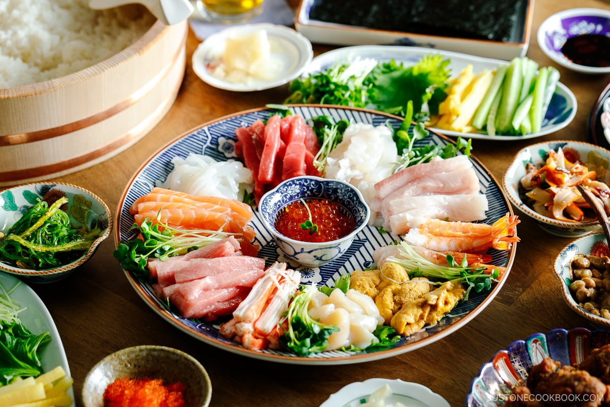 A temaki hand roll sushi spread on the dining table, consisting of plates and bowls containing sushi rice, sashimi, vegetable fillings, and nori seaweed.