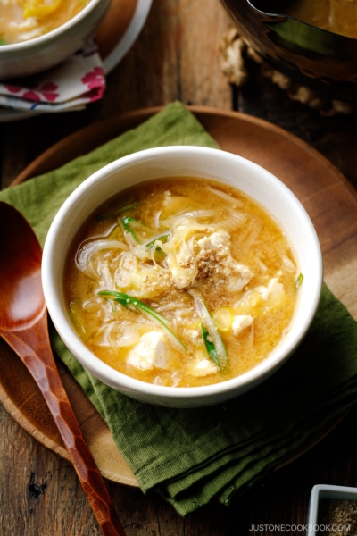 A white ceramic bowl containing Miso Soup with enoki mushrooms, tofu, and Japanese long green onion, topped with ground sesame seeds.