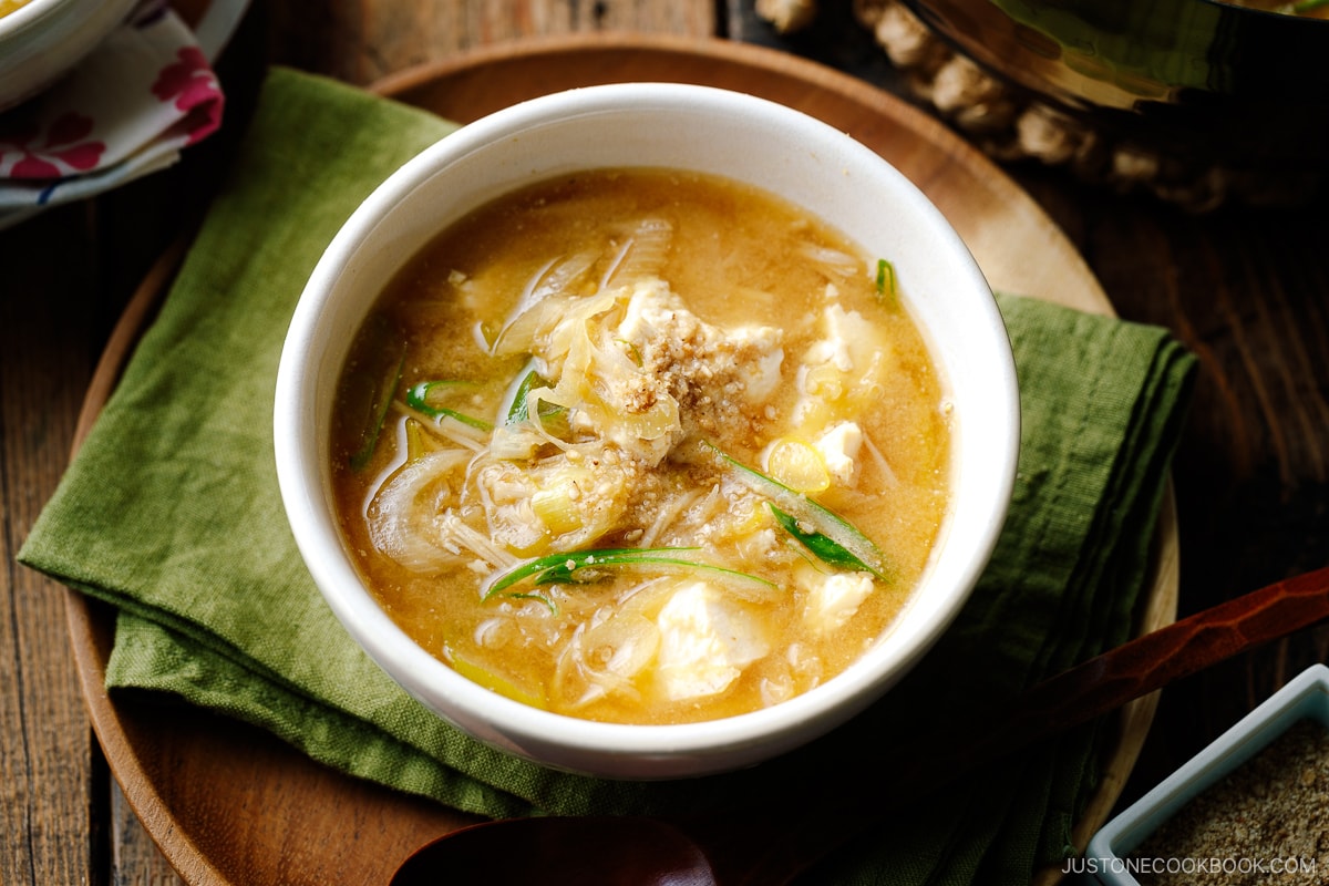 A white ceramic bowl containing Miso Soup with enoki mushrooms, tofu, and Japanese long green onion, topped with ground sesame seeds.