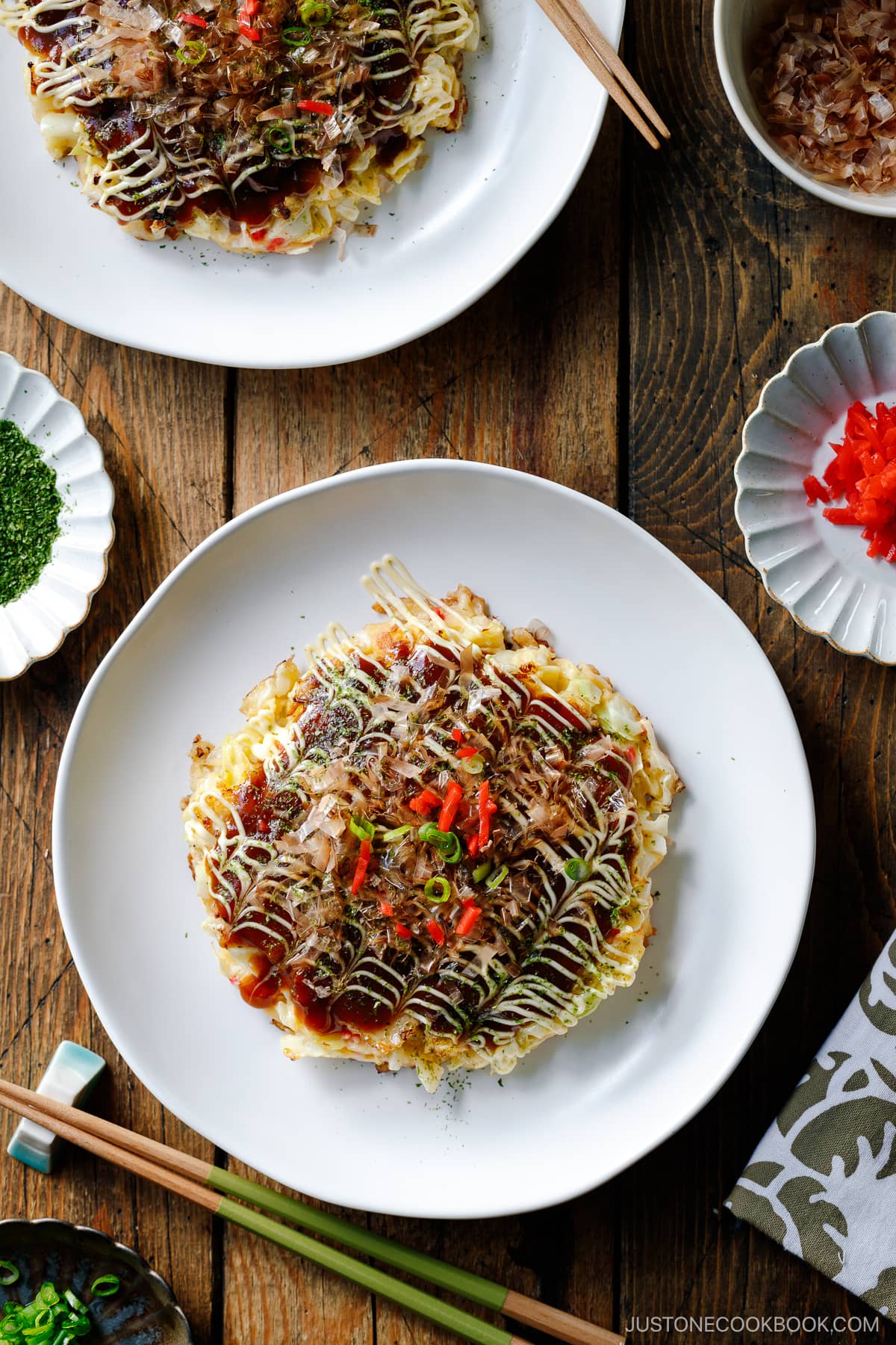 A white ceramic plate containing Okonomiyaki, a savory Japanese cabbage pancake topped with okonomi sauce, Kewpie mayo, and bonito flakes.