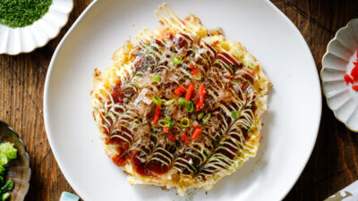 A white ceramic plate containing Okonomiyaki, a savory Japanese cabbage pancake topped with okonomi sauce, Kewpie mayo, and bonito flakes.