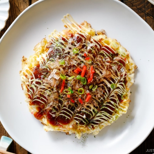 A white ceramic plate containing Okonomiyaki, a savory Japanese cabbage pancake topped with okonomi sauce, Kewpie mayo, and bonito flakes.