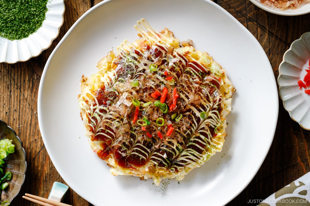 A white ceramic plate containing Okonomiyaki, a savory Japanese cabbage pancake topped with okonomi sauce, Kewpie mayo, and bonito flakes.