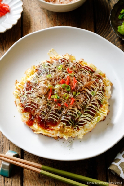 A white ceramic plate containing Okonomiyaki, a savory Japanese cabbage pancake topped with okonomi sauce, Kewpie mayo, and bonito flakes.