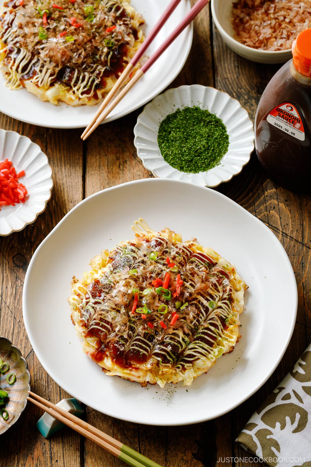 A white ceramic plate containing Okonomiyaki, a savory Japanese cabbage pancake topped with okonomi sauce, Kewpie mayo, and bonito flakes.