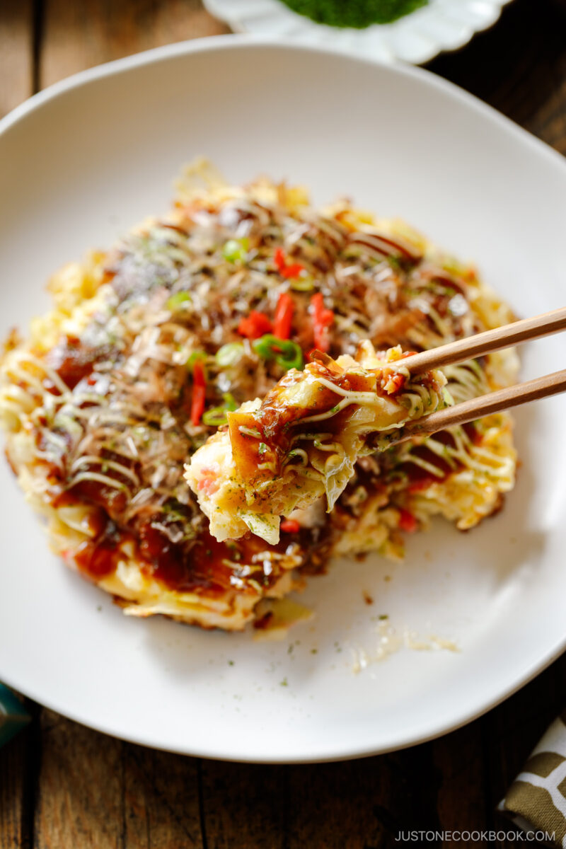 A white ceramic plate containing Okonomiyaki, a savory Japanese cabbage pancake topped with okonomi sauce, Kewpie mayo, and bonito flakes.