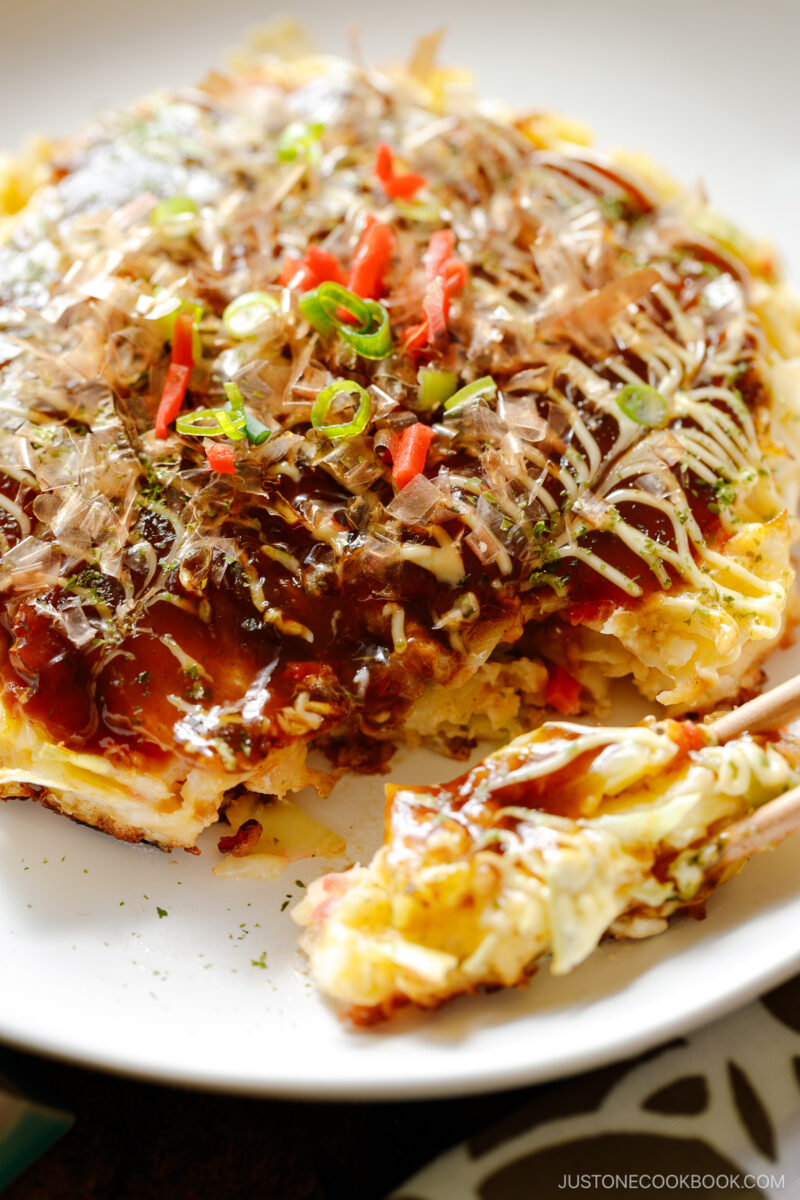 A white ceramic plate containing Okonomiyaki, a savory Japanese cabbage pancake topped with okonomi sauce, Kewpie mayo, and bonito flakes.