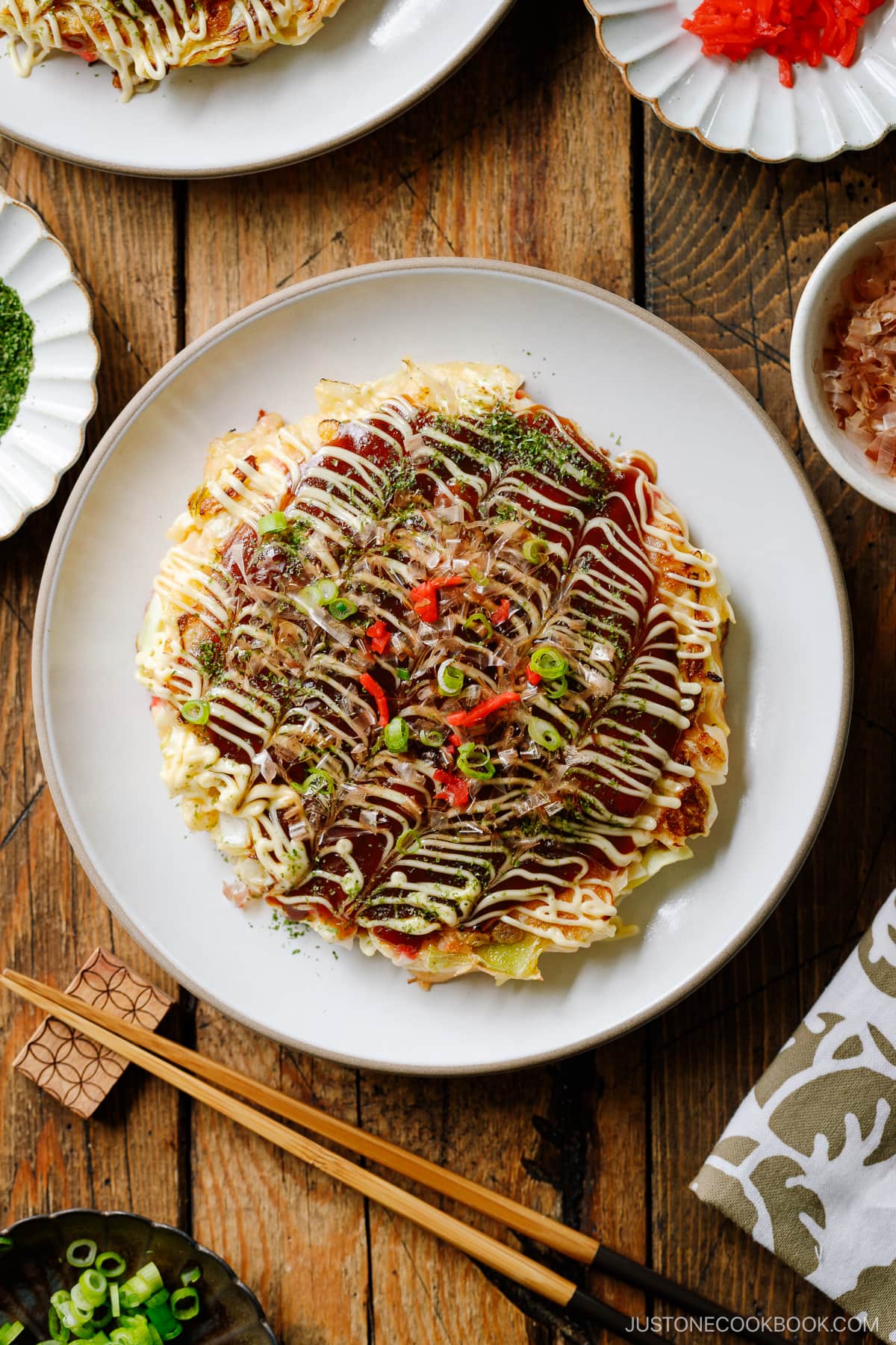 A white ceramic plate containing Okonomiyaki, a savory Japanese cabbage pancake topped with okonomi sauce, Kewpie mayo, and bonito flakes.