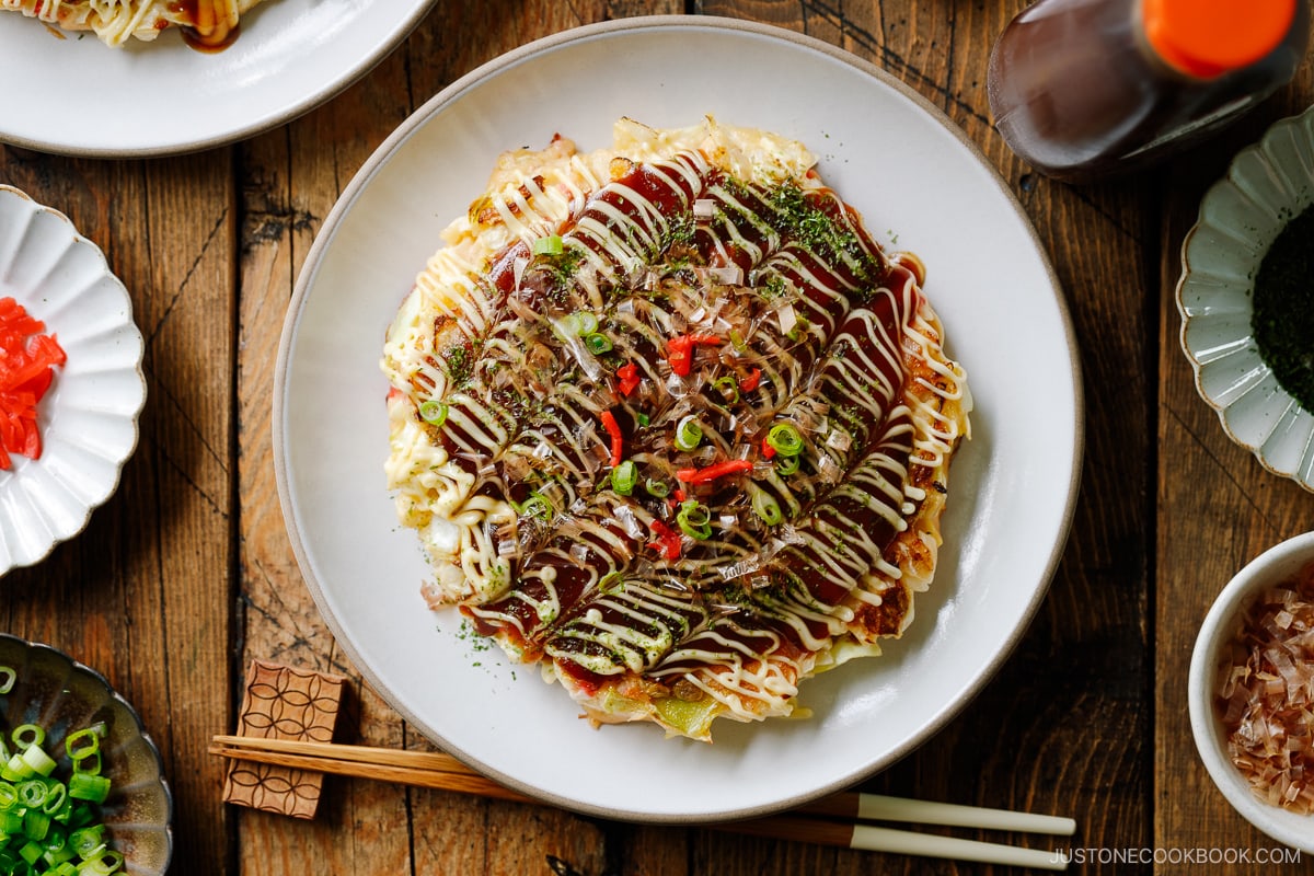 A white ceramic plate containing Okonomiyaki, a savory Japanese cabbage pancake topped with okonomi sauce, Kewpie mayo, and bonito flakes.