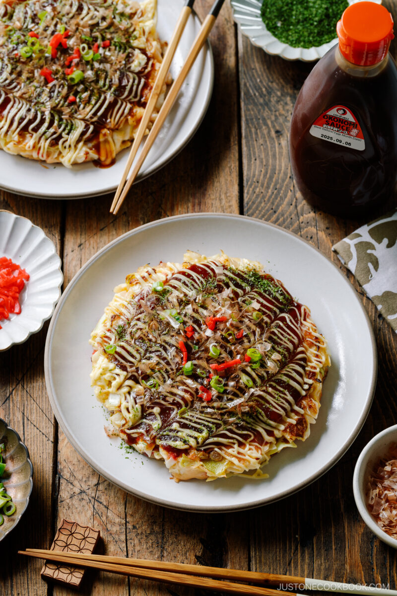 A white ceramic plate containing Okonomiyaki, a savory Japanese cabbage pancake topped with okonomi sauce, Kewpie mayo, and bonito flakes.