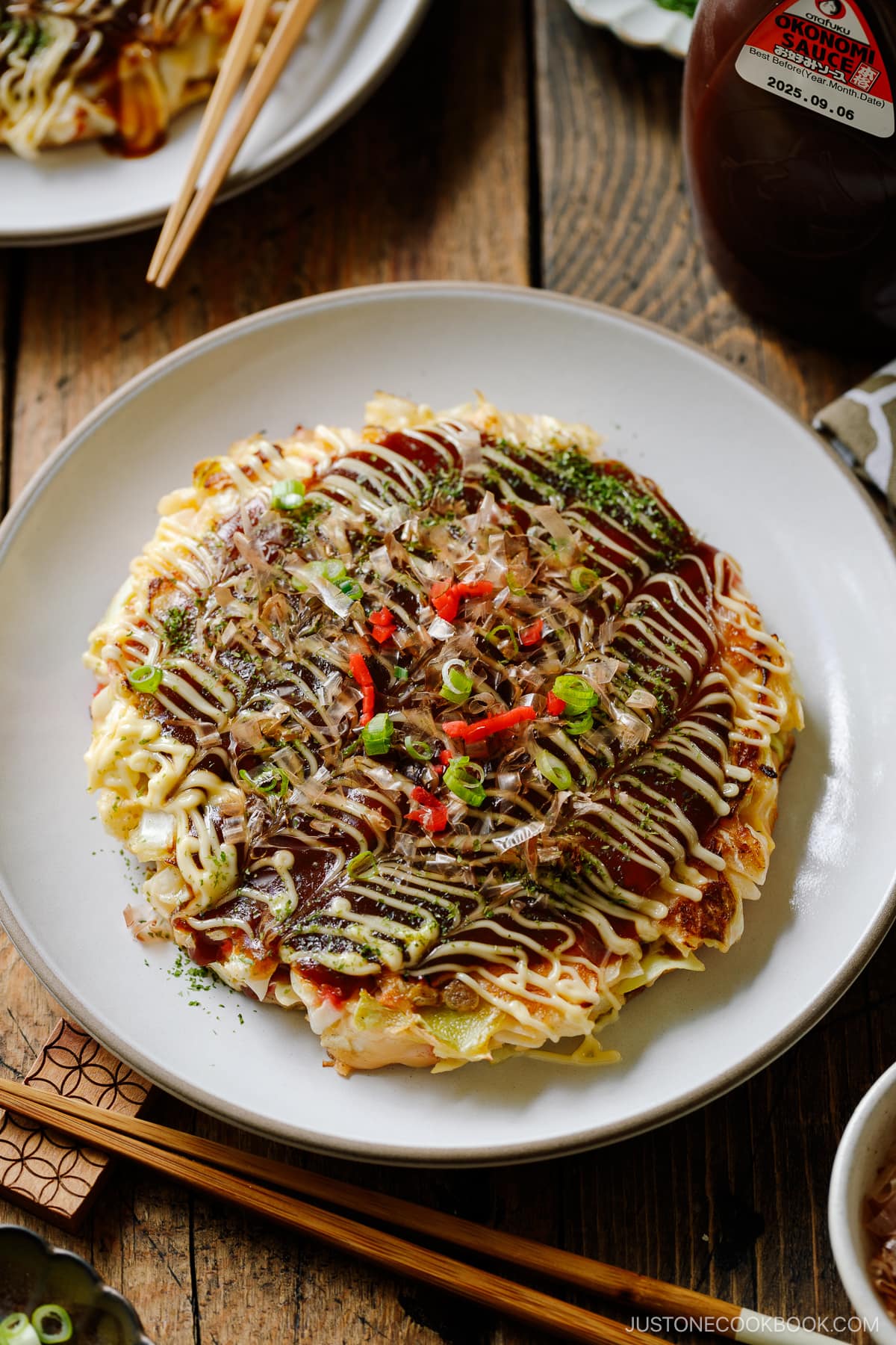 A white ceramic plate containing Okonomiyaki, a savory Japanese cabbage pancake topped with okonomi sauce, Kewpie mayo, and bonito flakes.