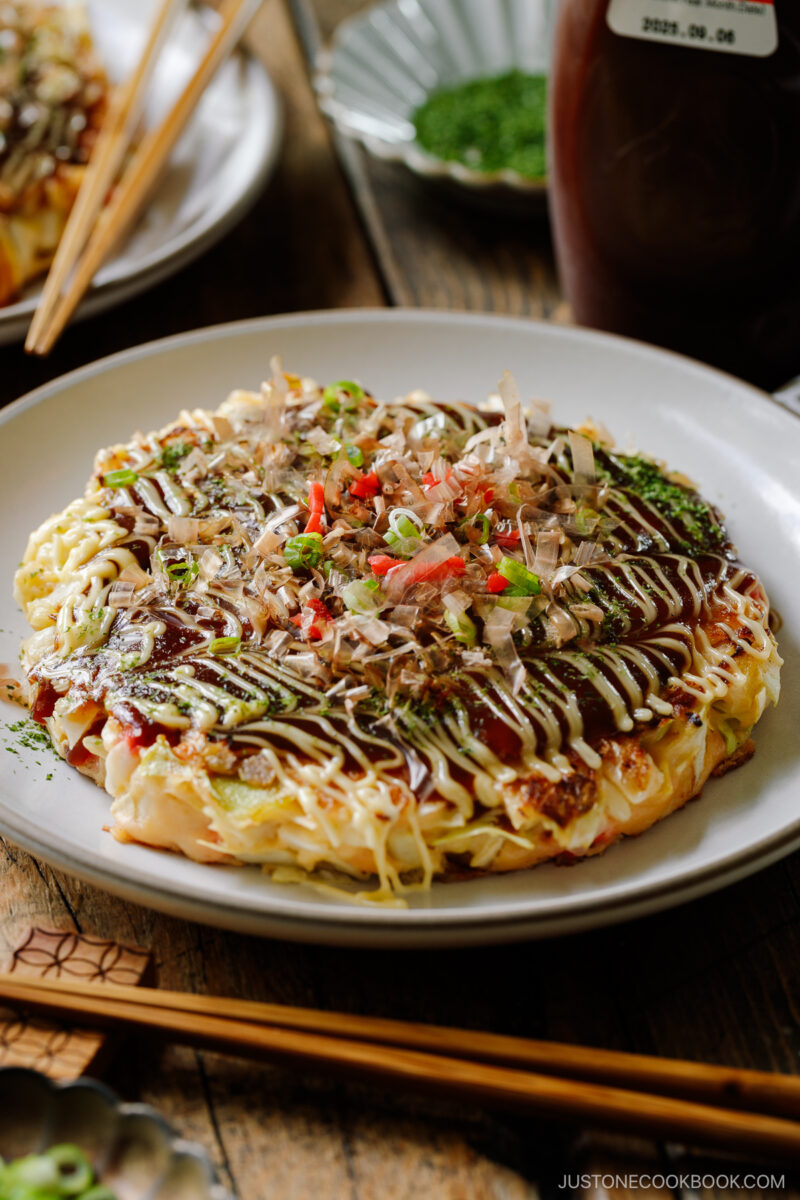 A white ceramic plate containing Okonomiyaki, a savory Japanese cabbage pancake topped with okonomi sauce, Kewpie mayo, and bonito flakes.