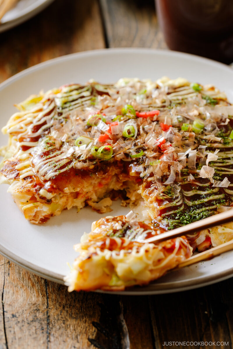 A white ceramic plate containing Okonomiyaki, a savory Japanese cabbage pancake topped with okonomi sauce, Kewpie mayo, and bonito flakes.