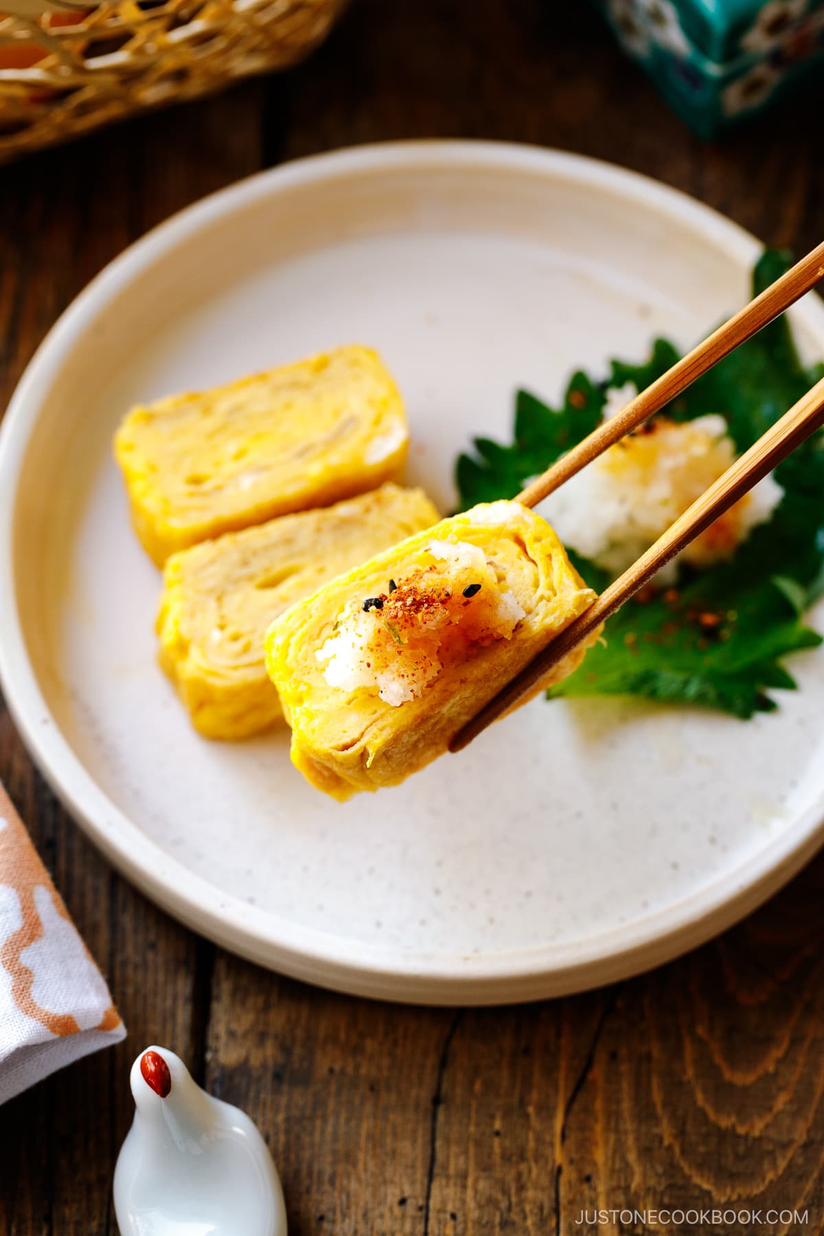 A round white plate containing Japanese rolled omelette, Tamagoyaki, along with shiso leaf and greated daikon drizzled with soy sauce and sprinkled with shichimi togarashi.