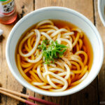 Japanese bowls containing Classic Udon Noodle Soup called Kake Udon or Su Udon, topped with sliced green onion and shichimi togarashi (Japanese seven spice).