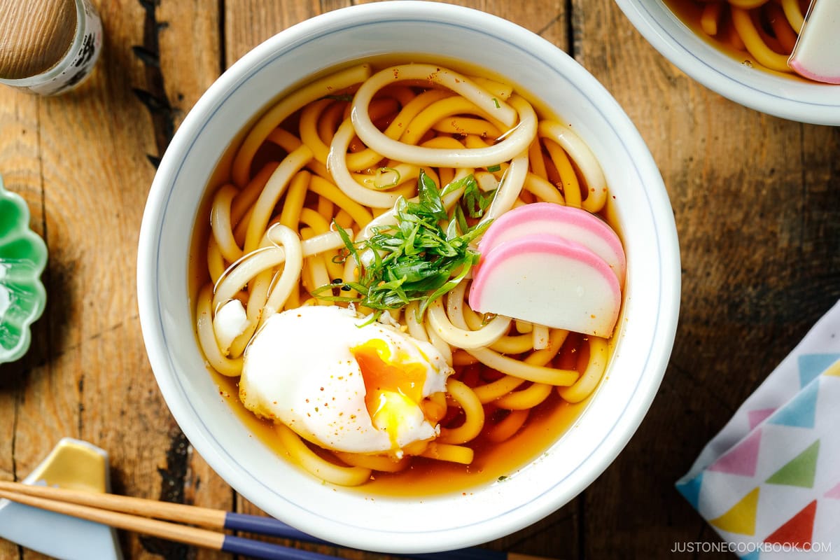 Japanese bowls containing Classic Udon Noodle Soup called Kake Udon or Su Udon, topped with sliced green onion, onsen tamago (poached egg), and fish cake slices.