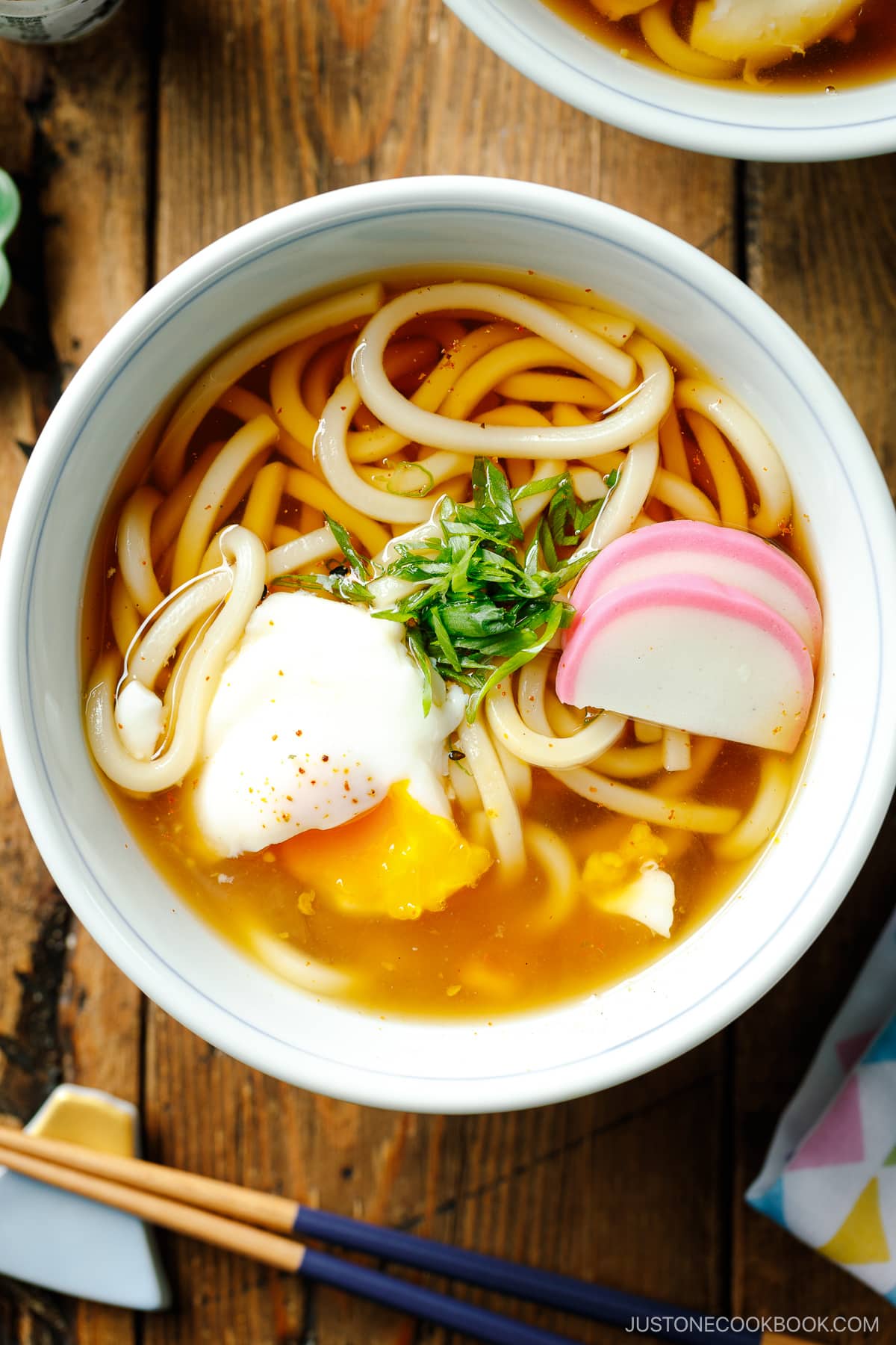 Japanese bowls containing Classic Udon Noodle Soup called Kake Udon or Su Udon, topped with sliced green onion, onsen tamago (poached egg), and fish cake slices.
