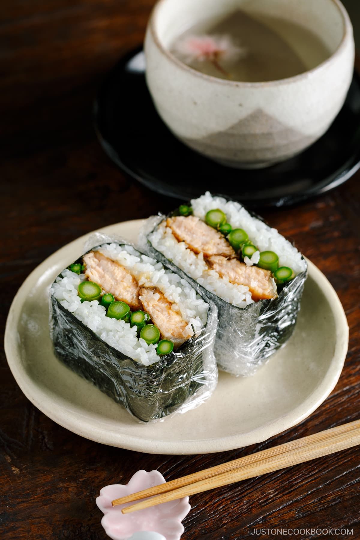 A white p late containing Teriyaki Salmon Onigirazu wrapped in plastic and showing the cross section.