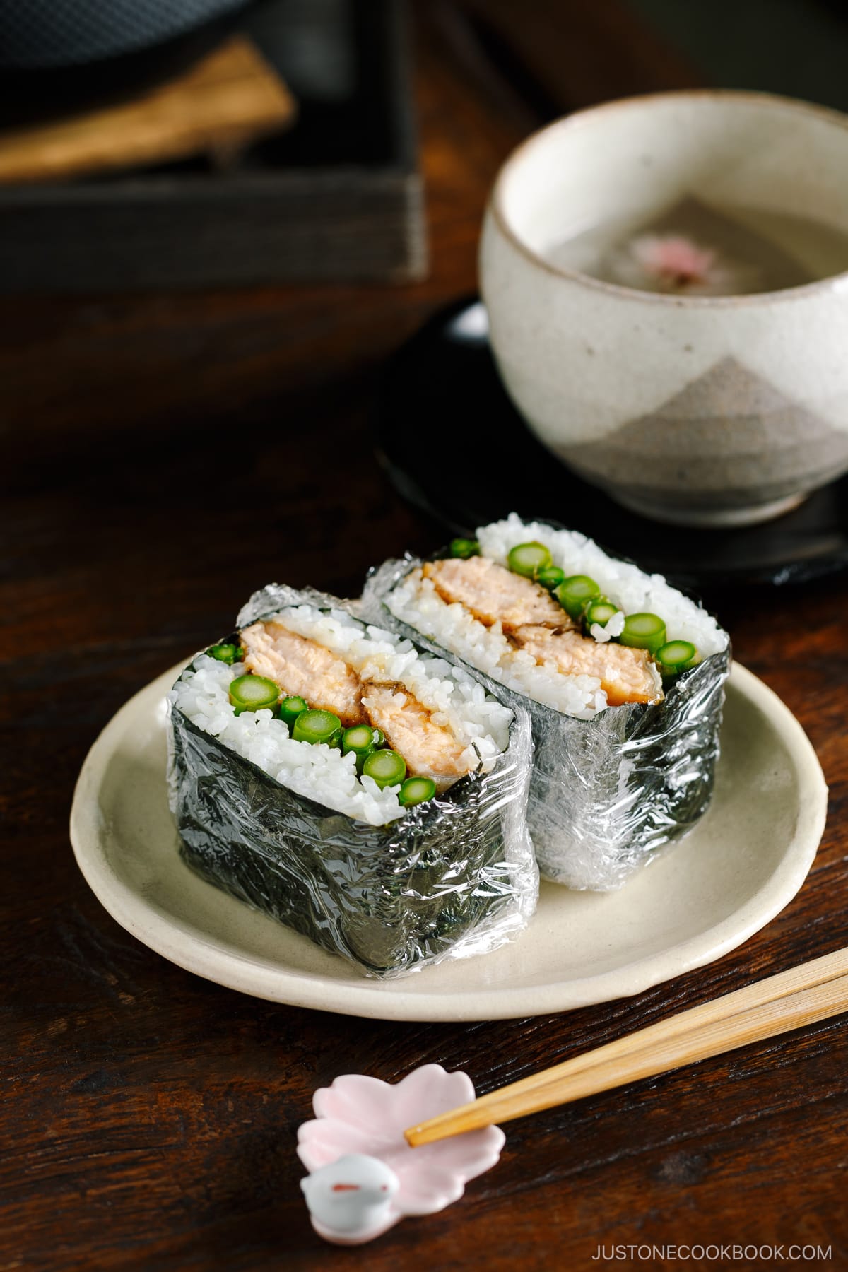 A white p late containing Teriyaki Salmon Onigirazu wrapped in plastic and showing the cross section.