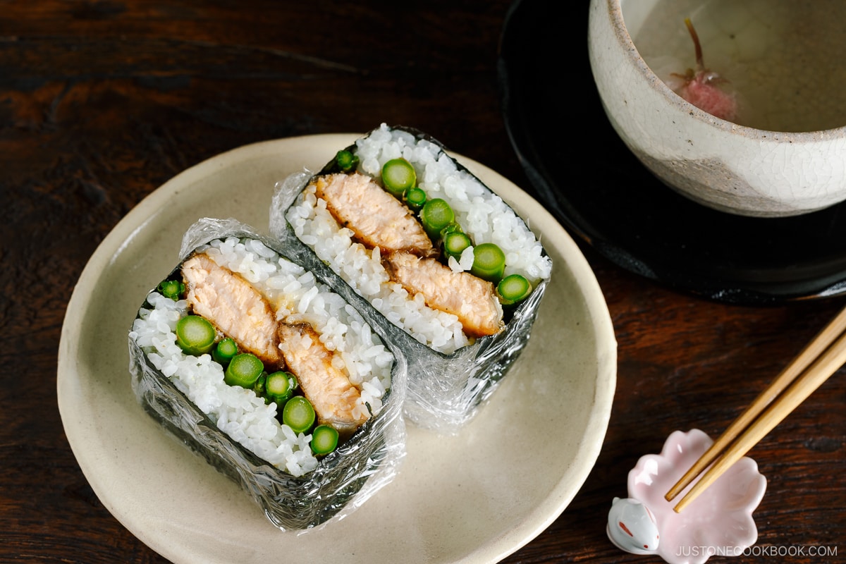 A white p late containing Teriyaki Salmon Onigirazu wrapped in plastic and showing the cross section.