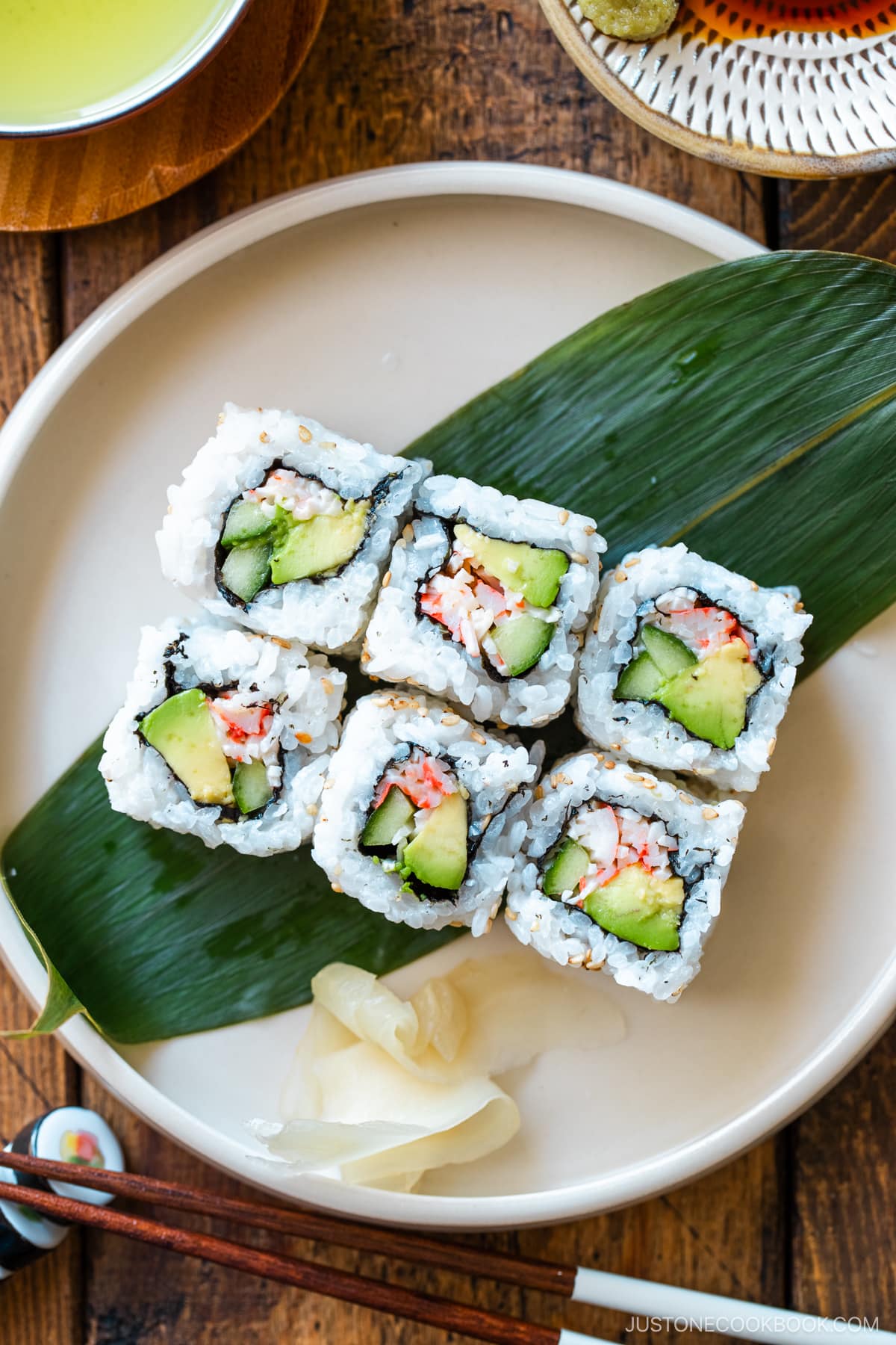 A round ceramic plate containing six pieces of California rolls over a bamboo leaf along with sushi ginger.