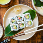 Round ceramic plates containing six pieces of California rolls over a bamboo leaf along with sushi ginger.