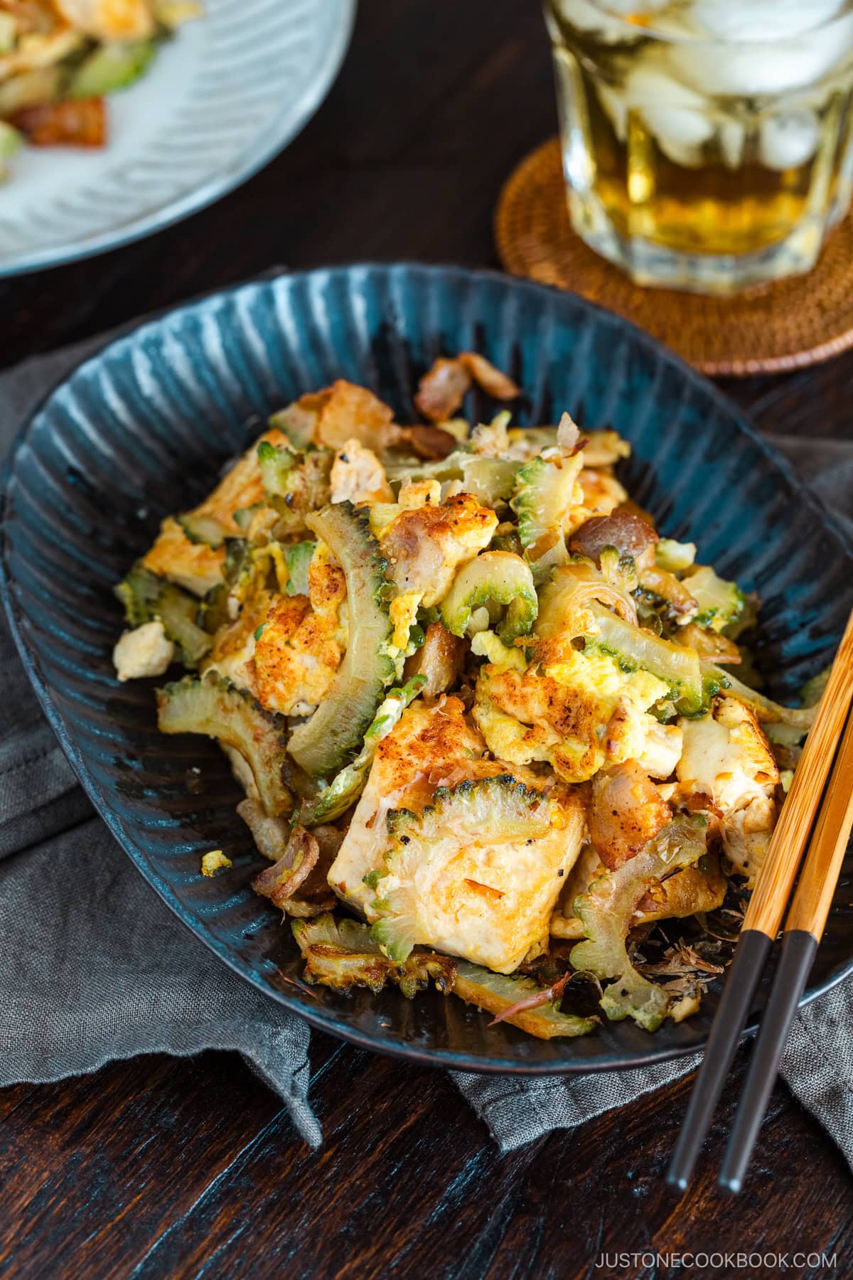 A dark oval plate containing Goya Champuru, a bitter melon stir-fry with tofu, egg, and pork belly.