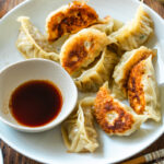 A round plate containing gyoza (Japanese potstickers or pan-fried dumplings) with a small plate of dipping sauce made with soy sauce, vinegar, and Japanese chili oil.