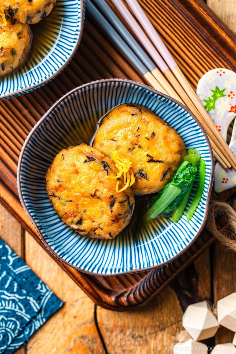 A blue and white bowl containing Simmered Ganmodoki topped with yuzu zest and served with dashi soy broth and komatsuna greens.