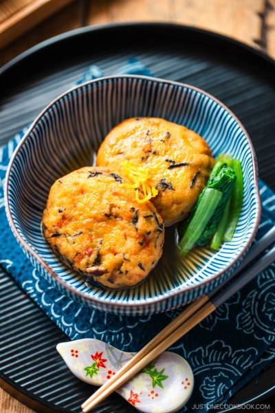 A blue and white bowl containing Simmered Ganmodoki topped with yuzu zest and served with dashi soy broth and komatsuna greens.