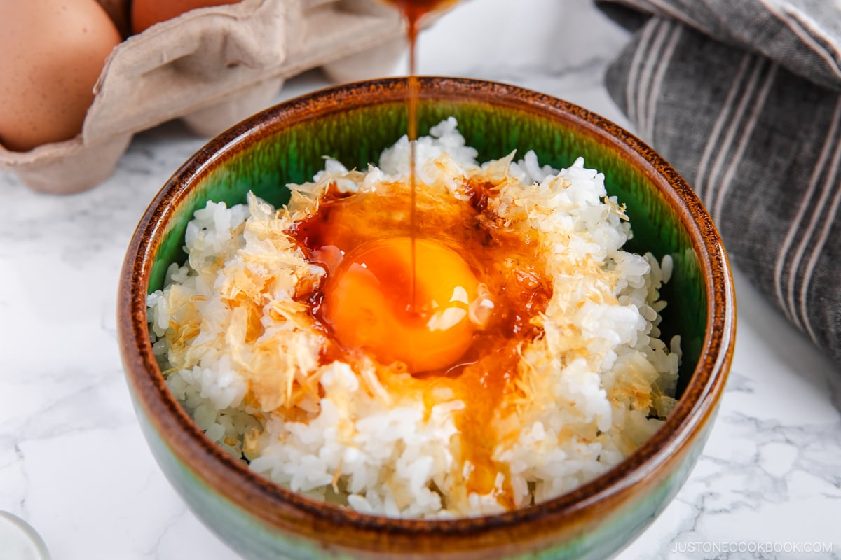 A bowl containing steamed rice, raw egg, sprinkled katsuobushi, and a drizzle of soy sauce.