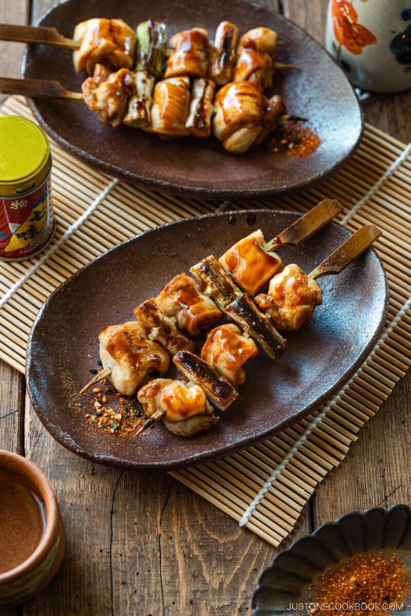 Oval rustic plates containing yakitori, Japanese grilled chicken and scallion skewers, coated with savory yakitori sauce and served with shichimi togarashi on the side.