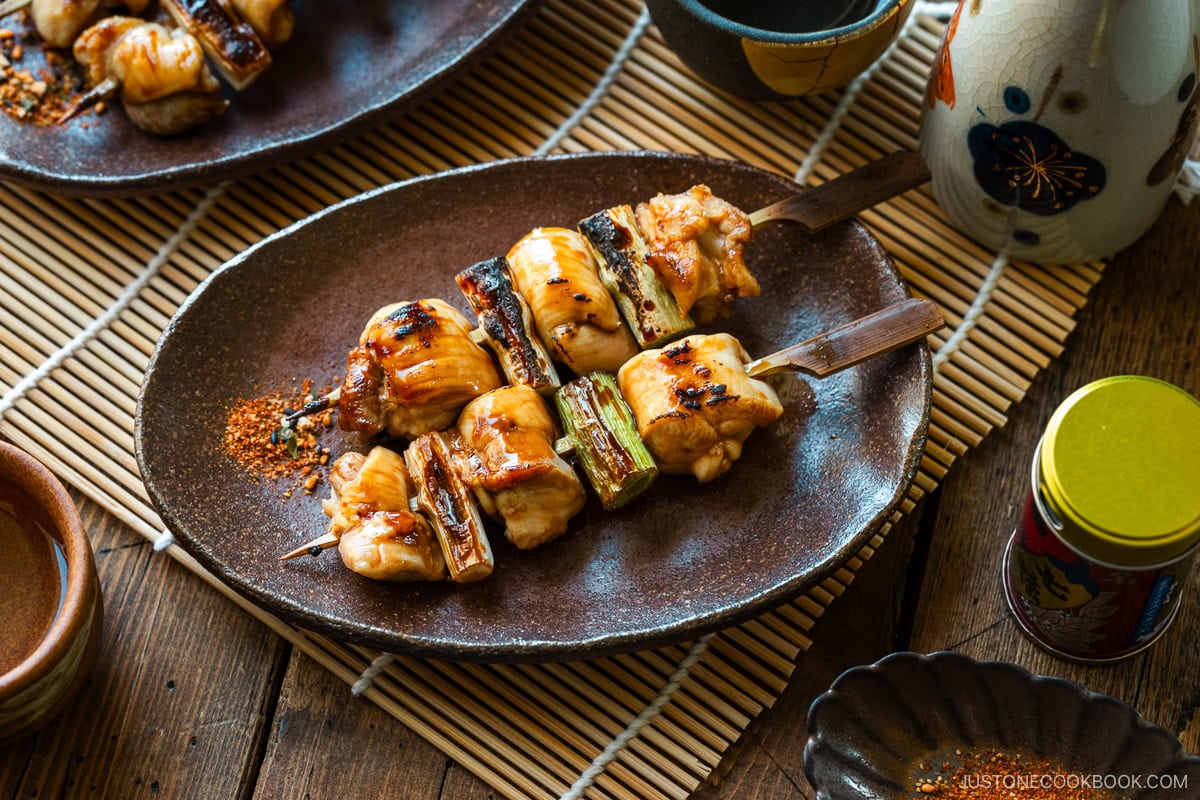 Oval rustic plates containing yakitori, Japanese grilled chicken and scallion skewers, coated with savory yakitori sauce and served with shichimi togarashi on the side.