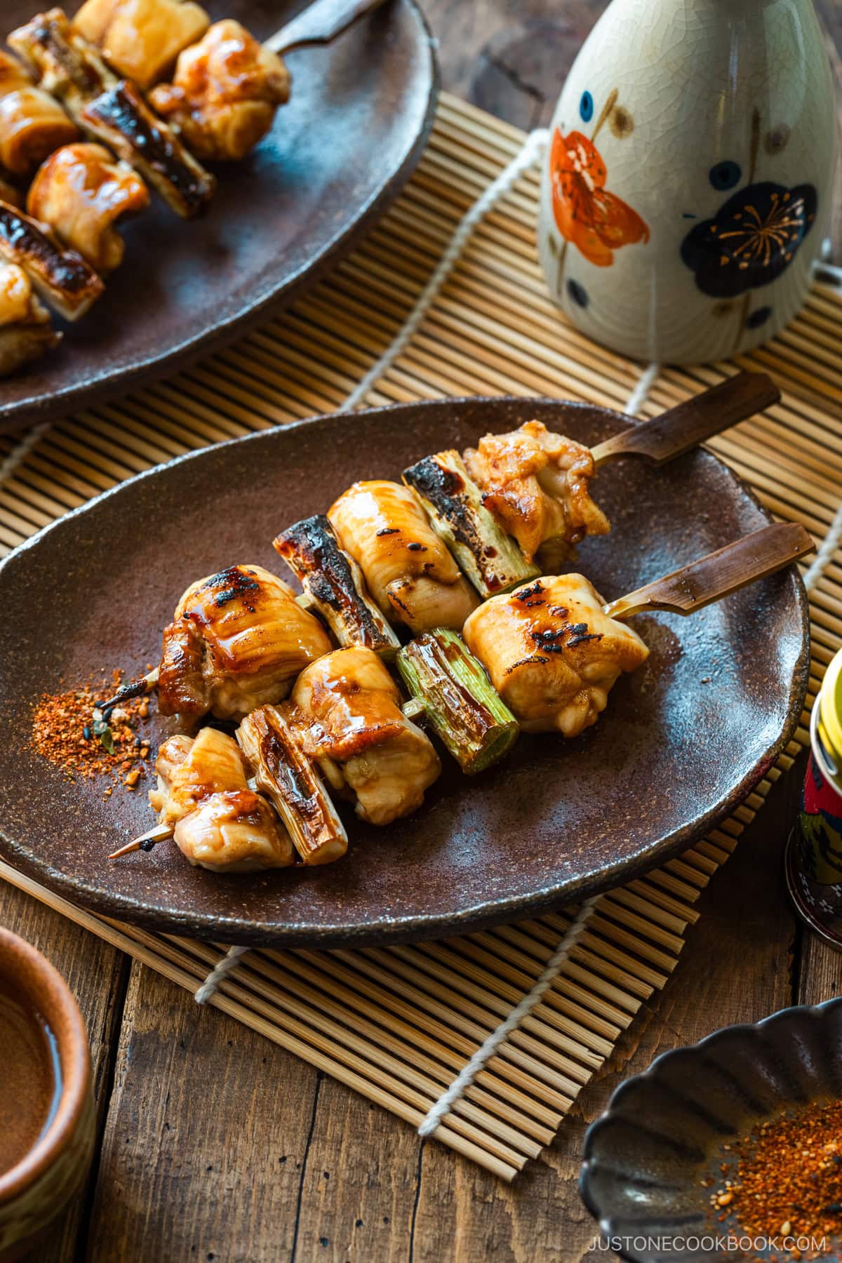 Oval rustic plates containing yakitori, Japanese grilled chicken and scallion skewers, coated with savory yakitori sauce and served with shichimi togarashi on the side.