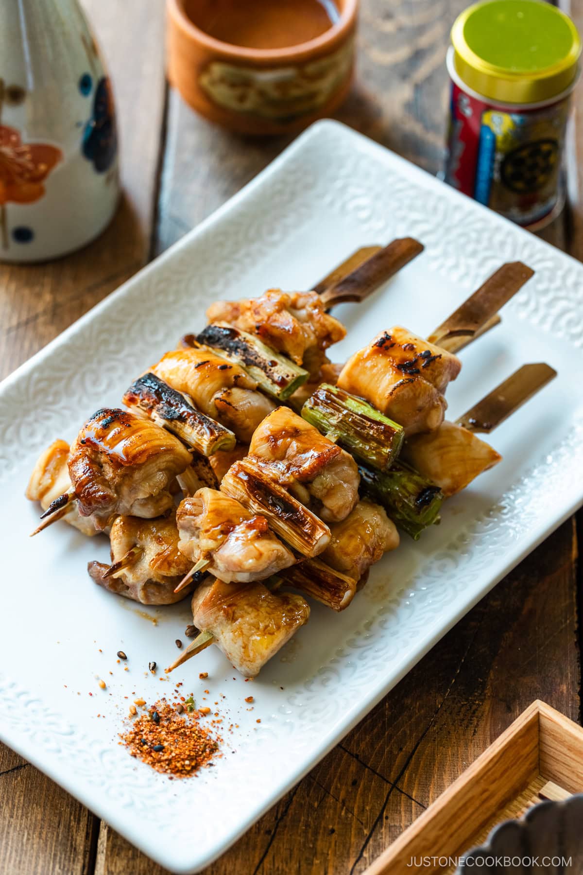 A white rectangular plate containing yakitori, Japanese grilled chicken and scallion skewers, coated with savory yakitori sauce and served with shichimi togarashi on the side.