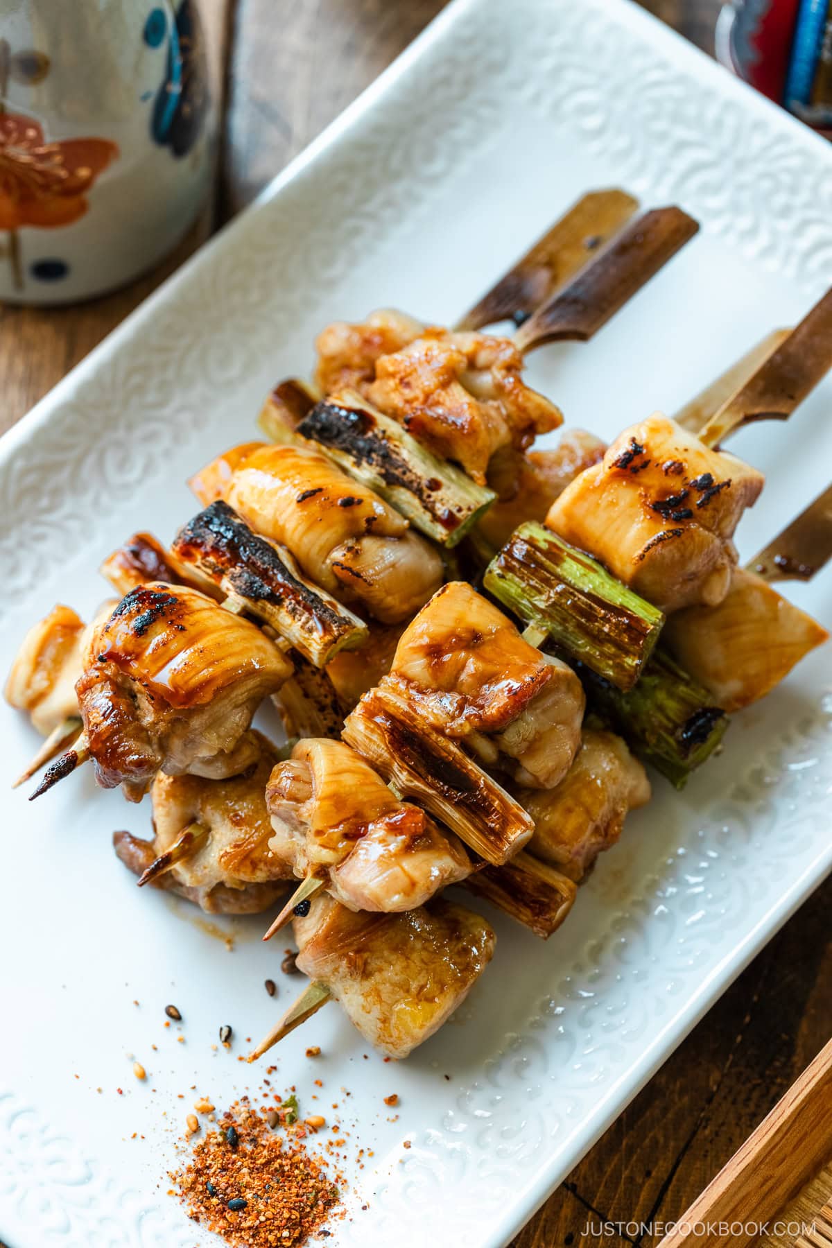 A white rectangular plate containing yakitori, Japanese grilled chicken and scallion skewers, coated with savory yakitori sauce and served with shichimi togarashi on the side.