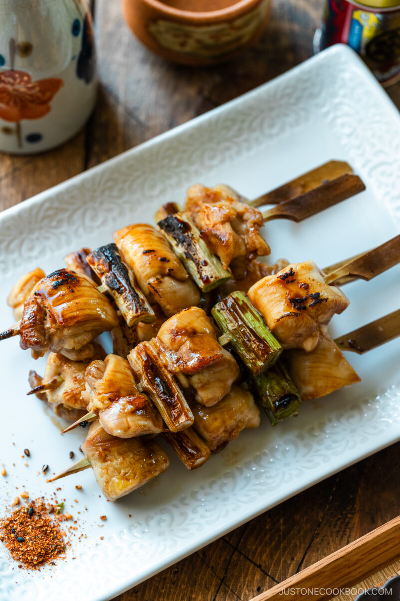 A white rectangular plate containing yakitori, Japanese grilled chicken and scallion skewers, coated with savory yakitori sauce and served with shichimi togarashi on the side.