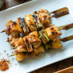 A white rectangular plate containing yakitori, Japanese grilled chicken and scallion skewers, coated with savory yakitori sauce and served with shichimi togarashi on the side.