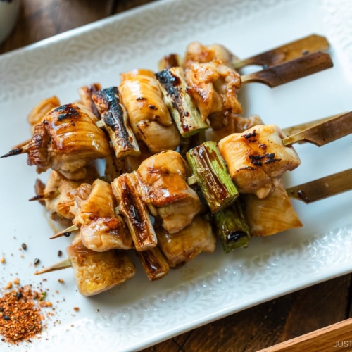 A white rectangular plate containing yakitori, Japanese grilled chicken and scallion skewers, coated with savory yakitori sauce and served with shichimi togarashi on the side.