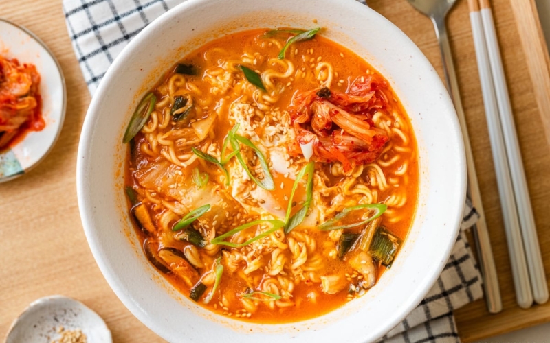 A white ramen bowl containing Korean ramen (ramyeon), topped with soft-cooked egg, kimchi, and green onions.